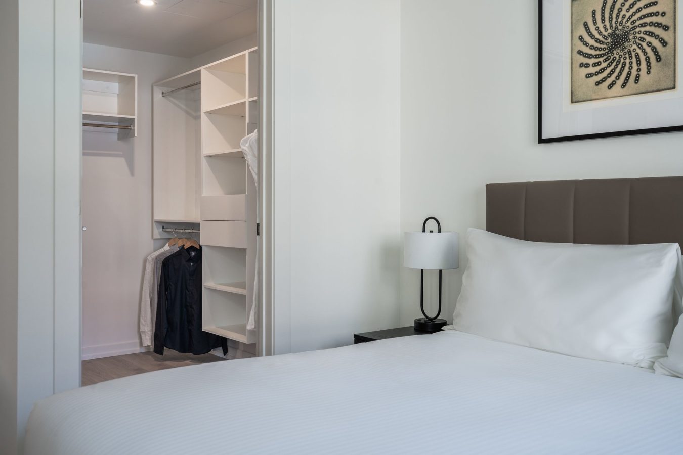 A neatly made bed with white sheets sits beside a nightstand and lamp. An open closet with hanging clothes and shelves is visible, and a framed artwork hangs on the white wall above the bed.
