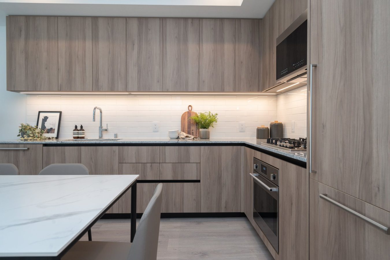 Modern kitchen with light wood cabinets, white tile backsplash, built-in stove and oven, under-cabinet lighting, and a marble dining table with gray chairs. Minimalist decor includes plants and framed art.