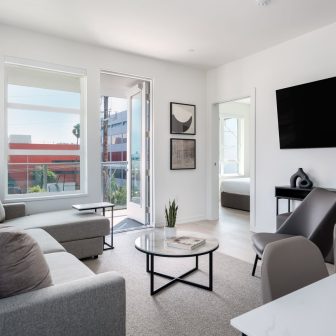 Modern living room with gray sofas, a round coffee table, wall-mounted TV, minimalist decor, large windows, and a door opening to a balcony. A bedroom can be seen through an open door in the background.