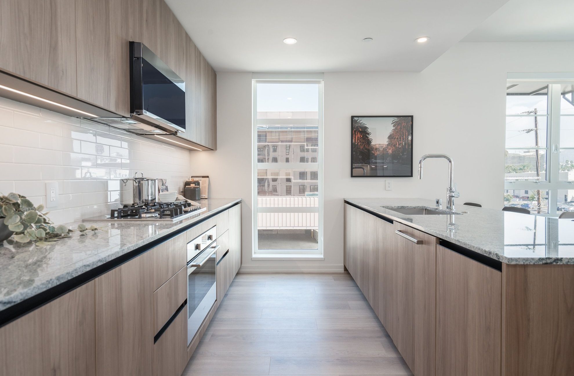 Modern kitchen with light wood cabinets, stainless steel appliances, a gas stove, and granite countertops. A tall window lets in natural light. A framed photo and a plant decorate the clean, minimalist space.