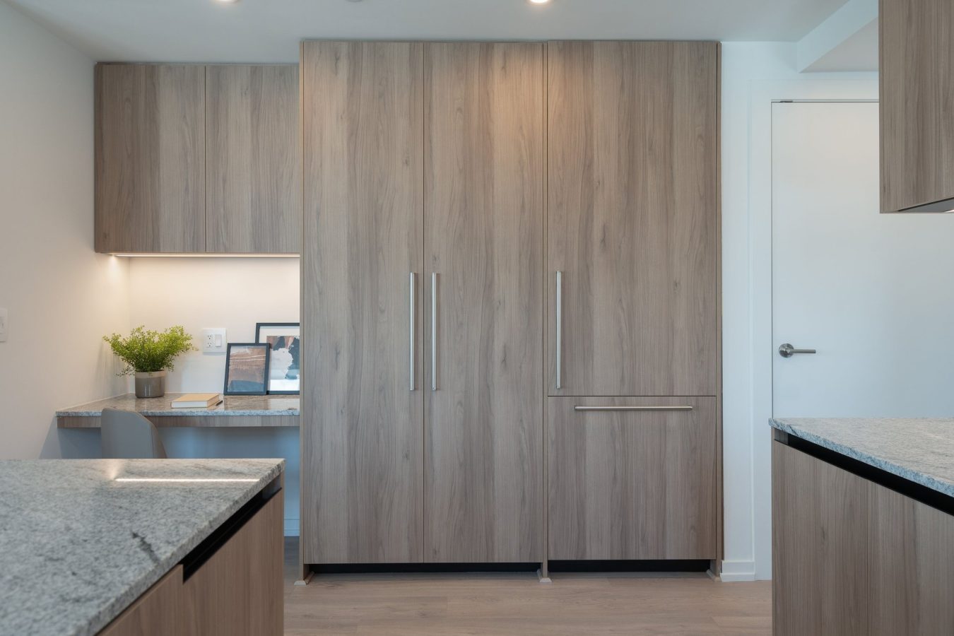 Minimalist kitchen with light wood cabinets, a built-in desk area with framed photos and a plant, gray stone countertops, and a closed white door on the right. The space is bright and modern.