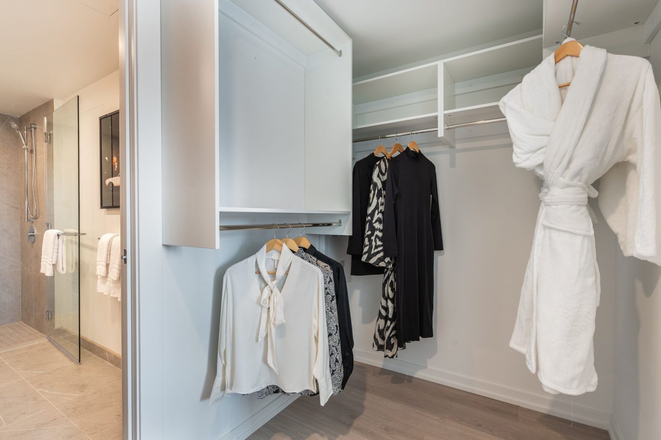 A modern walk-in closet with hanging clothes, including white blouses, a patterned dress, black dresses, and a white bathrobe. Shelves and wooden hangers are visible, with a bathroom in the background.