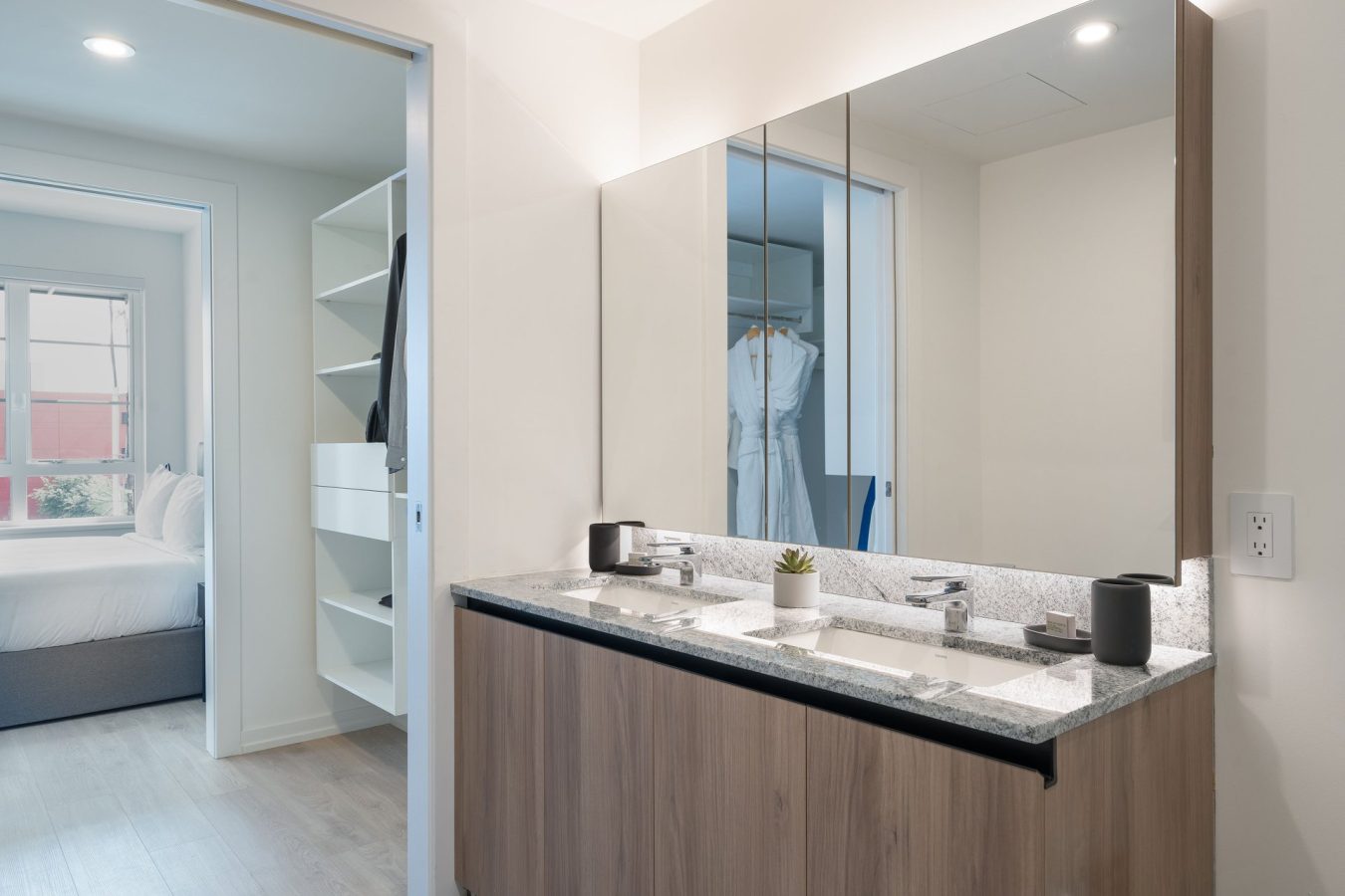 Modern bathroom with a double sink vanity, large mirror, and light wood cabinets. A walk-in closet with shelves and a bedroom with a window and bed are visible through open doorways.