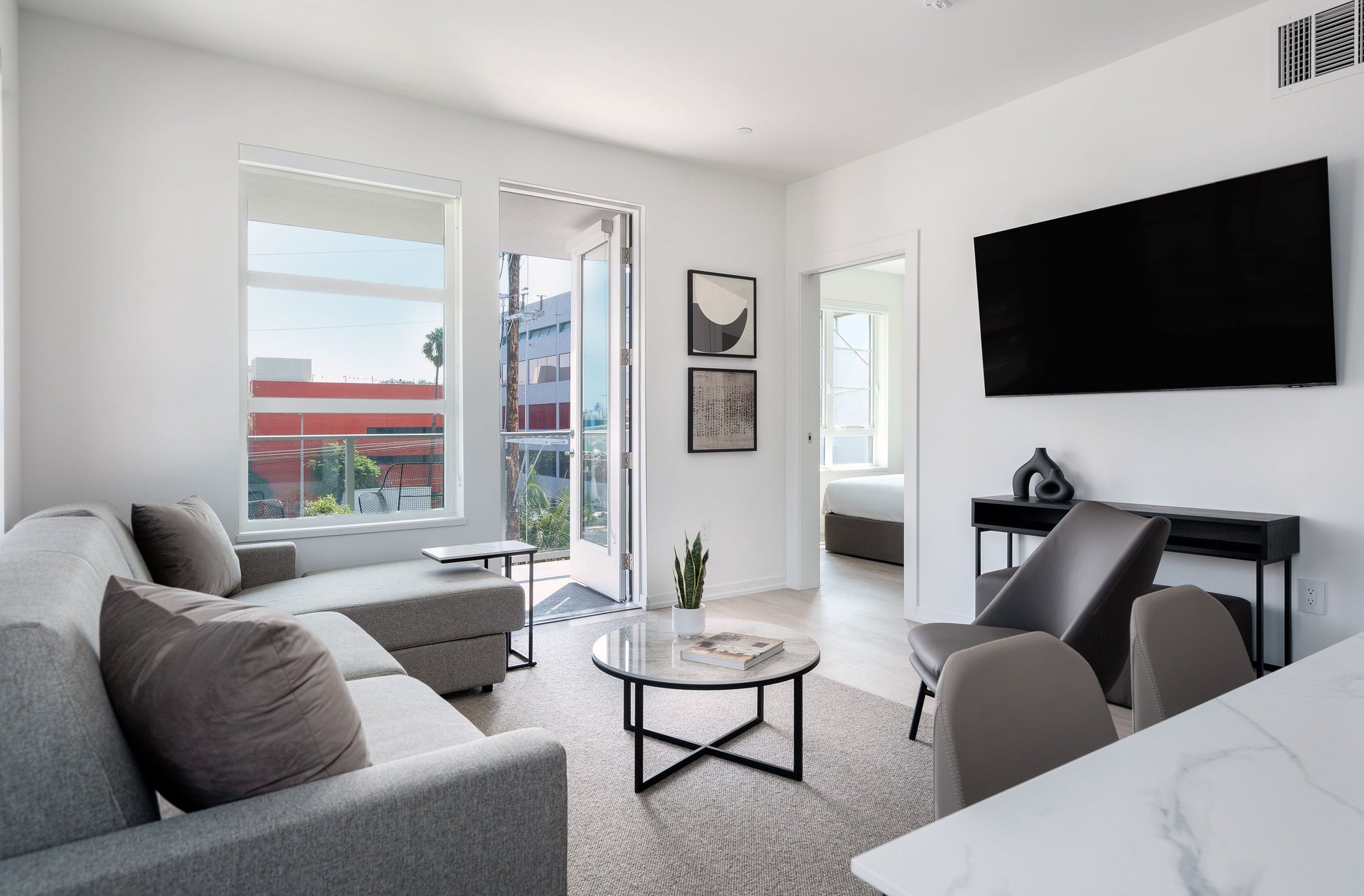 A modern, minimal living room with grey sofas, a round coffee table, wall-mounted TV, and large windows letting in natural light. A small plant sits on the table. A bedroom is visible through an open door.