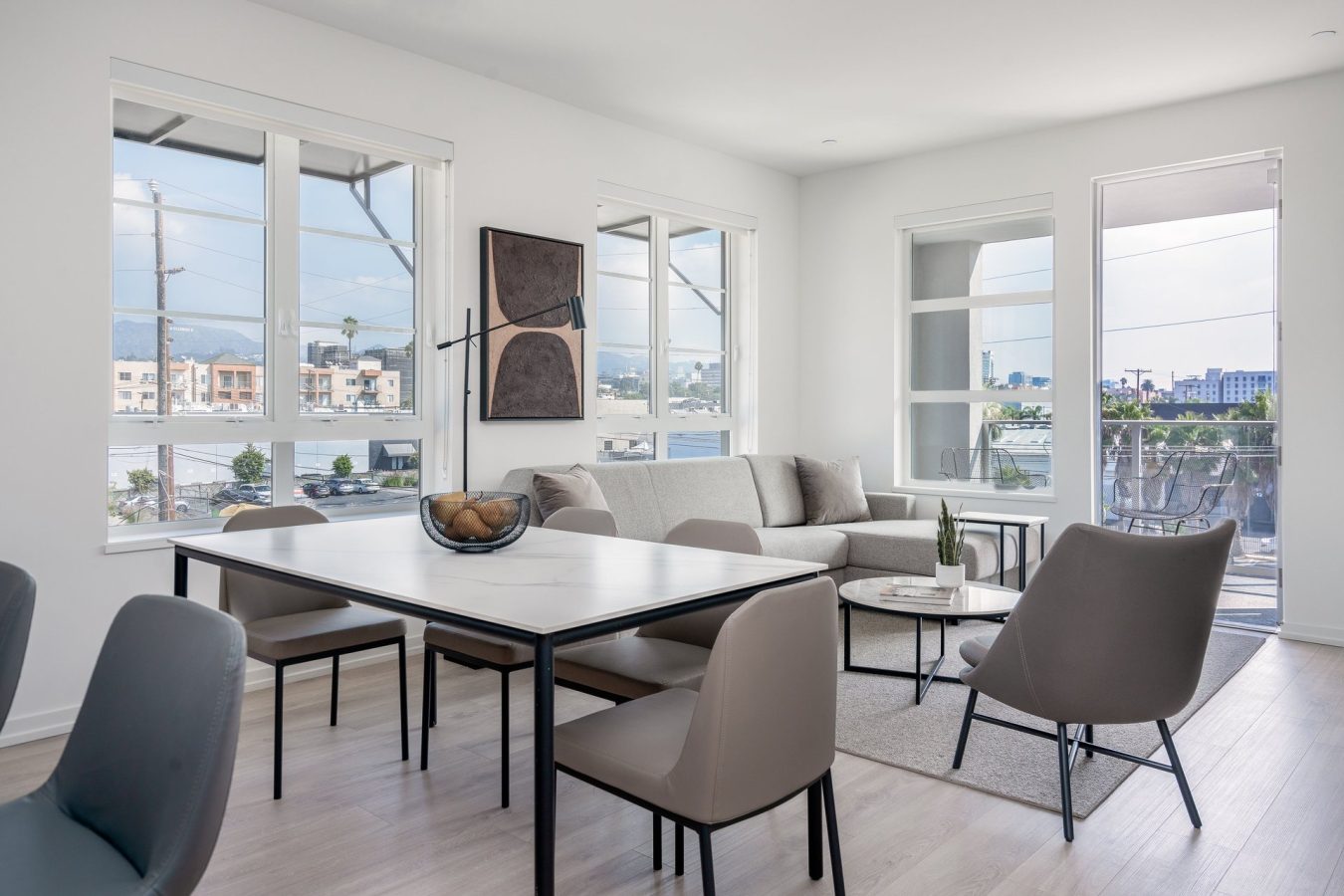 A modern living and dining area with large windows, a gray sofa, a marble dining table with beige chairs, and a coffee table. Natural light fills the room, highlighting minimalistic decor and outdoor views.