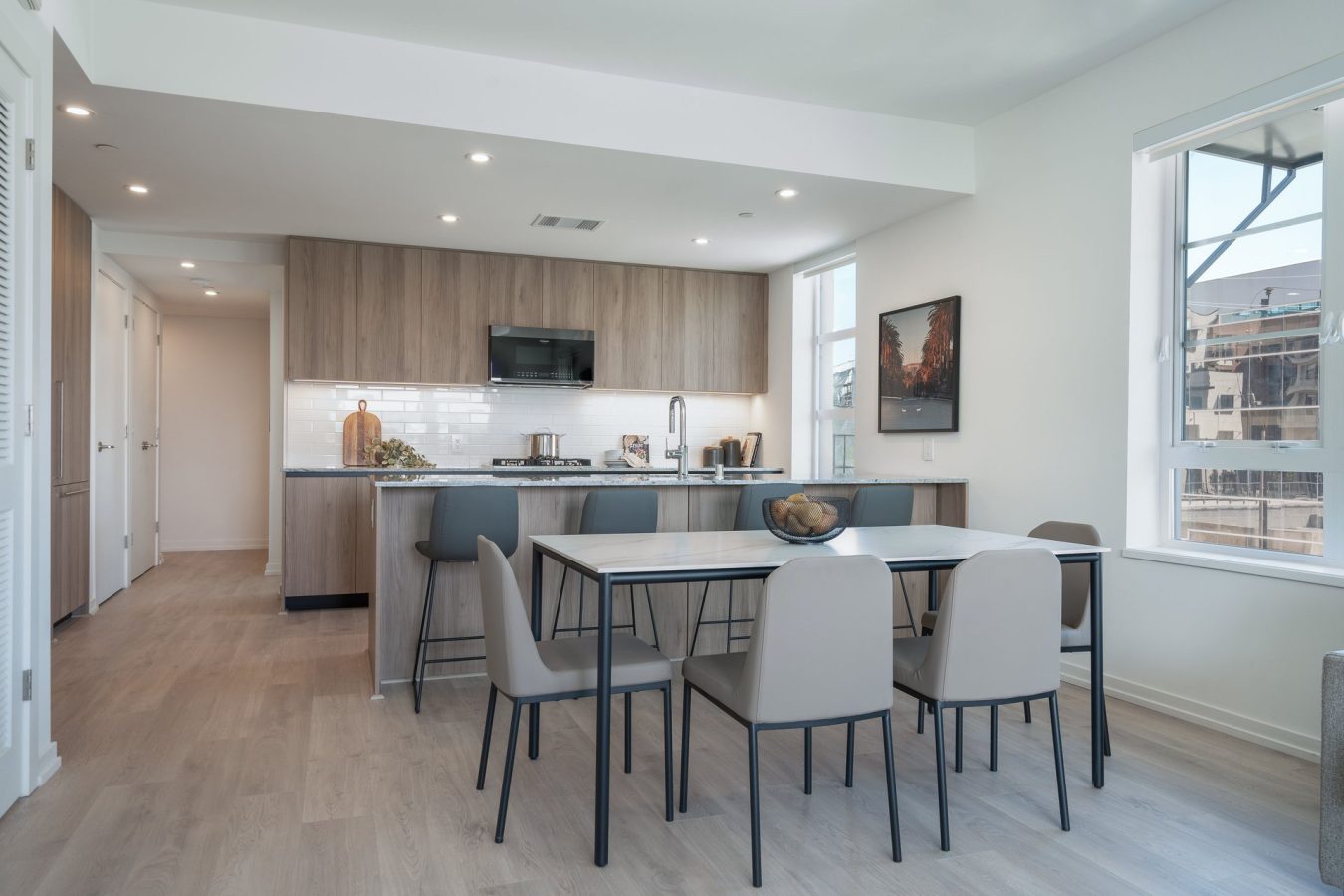 Modern kitchen and dining area with light wood floors, a rectangular dining table with six gray chairs, wood cabinets, and large windows letting in natural light. A bowl with fruit sits on the table.