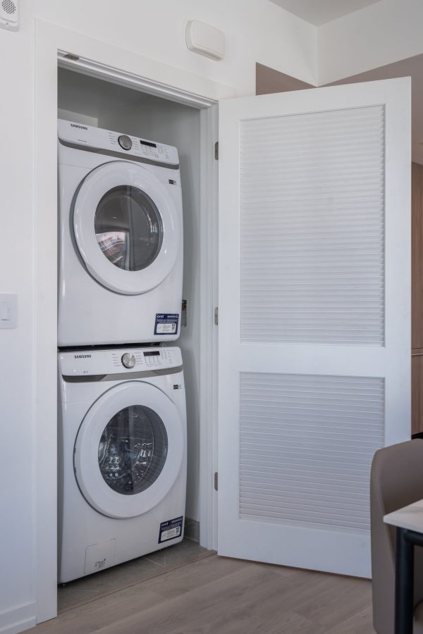 A stacked white washer and dryer set is installed in a small closet with a white louvered door partially open, in a modern laundry area with light wood flooring.