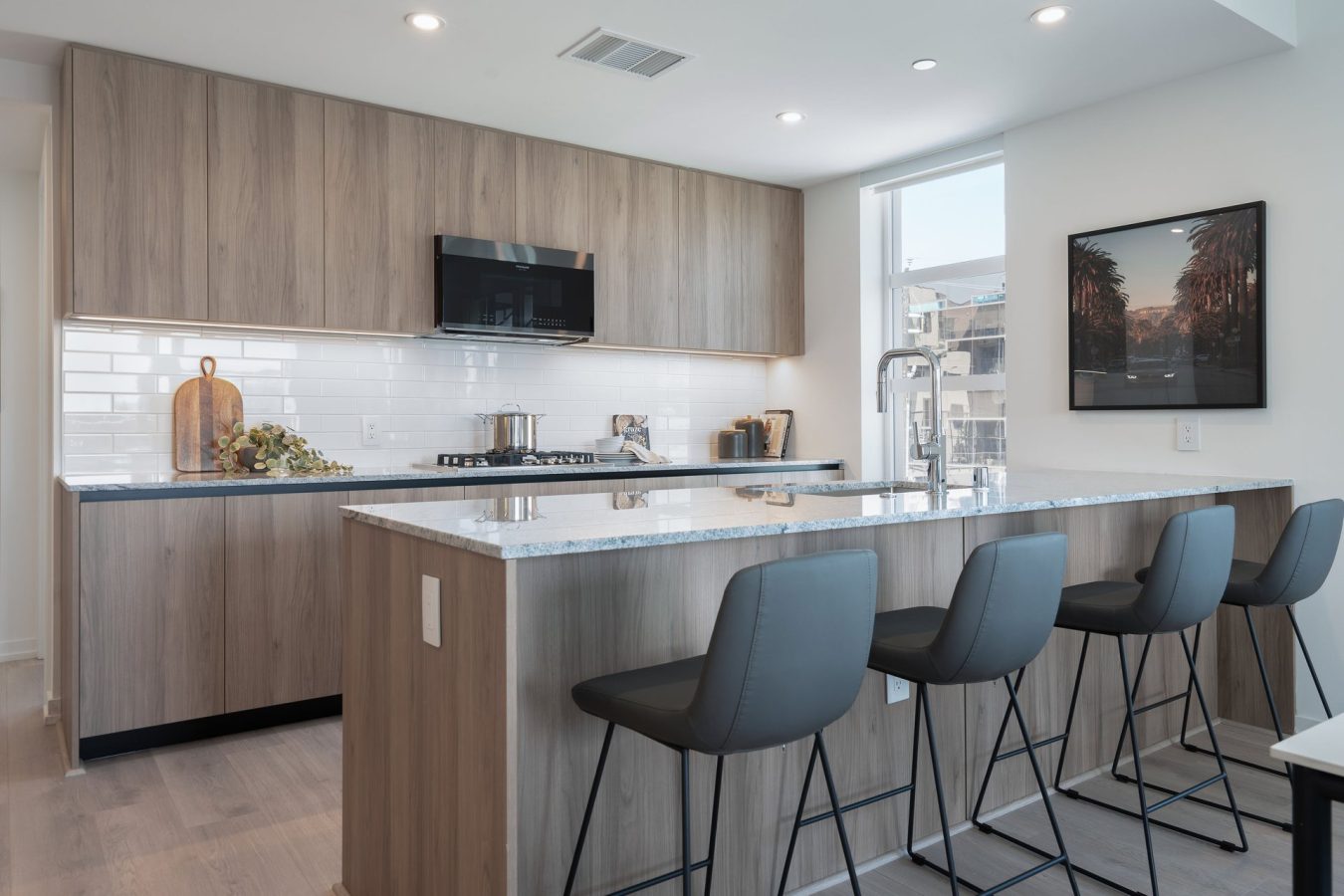 Modern kitchen with light wood cabinets, white subway tile backsplash, marble island with four gray barstools, stainless steel appliances, and a window letting in natural light. A framed picture hangs on the wall.