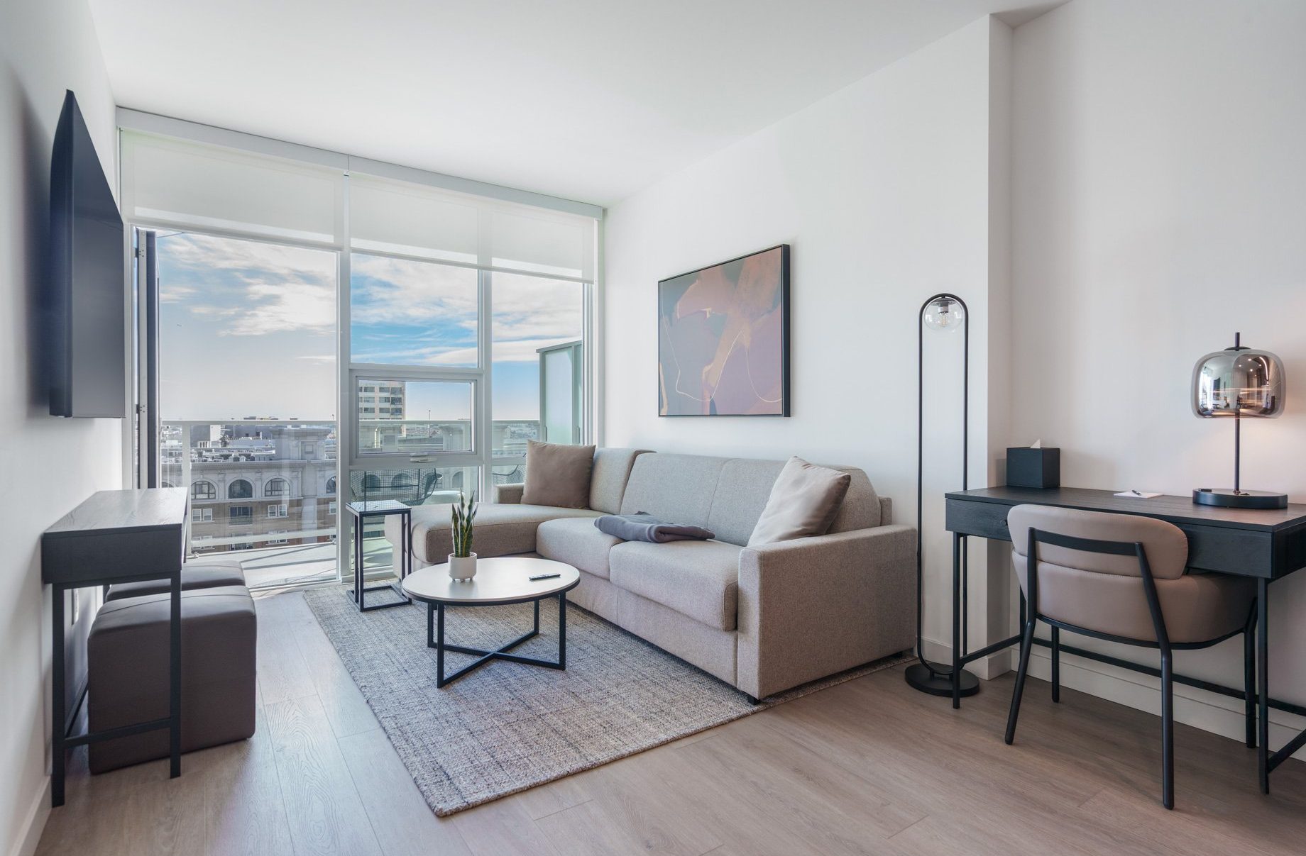 Modern living room with a gray sofa, round coffee table, desk with chair and lamp, flat-screen TV, abstract wall art, large windows with city view, and light wood flooring.
