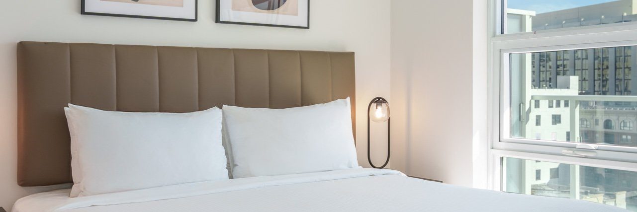 A neatly made bed with white bedding and pillows sits against a padded brown headboard. There are two framed artworks above the bed, a bedside lamp, and a large window showing buildings outside.