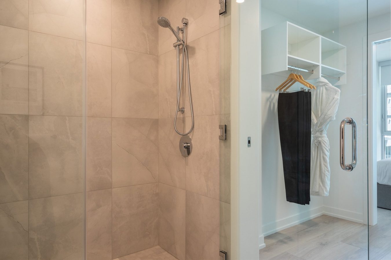 A modern bathroom with a glass-walled shower featuring beige tiles and a handheld showerhead. Adjacent is a walk-in closet with hanging shelves, a white bathrobe, and black pants on wooden hangers.