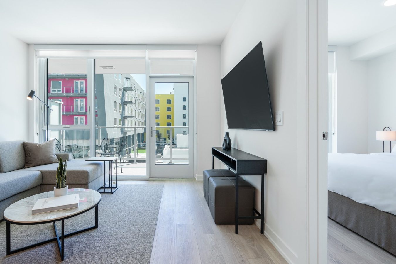 Modern apartment living room with a gray sofa, round marble coffee table, wall-mounted TV, and large windows opening to a balcony; bedroom visible through an open doorway.