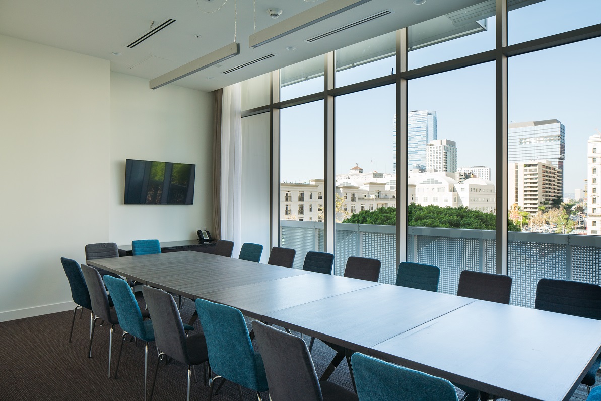 A modern conference room with a long table surrounded by blue chairs, a wall-mounted TV, large windows, and a city skyline view in the background.
