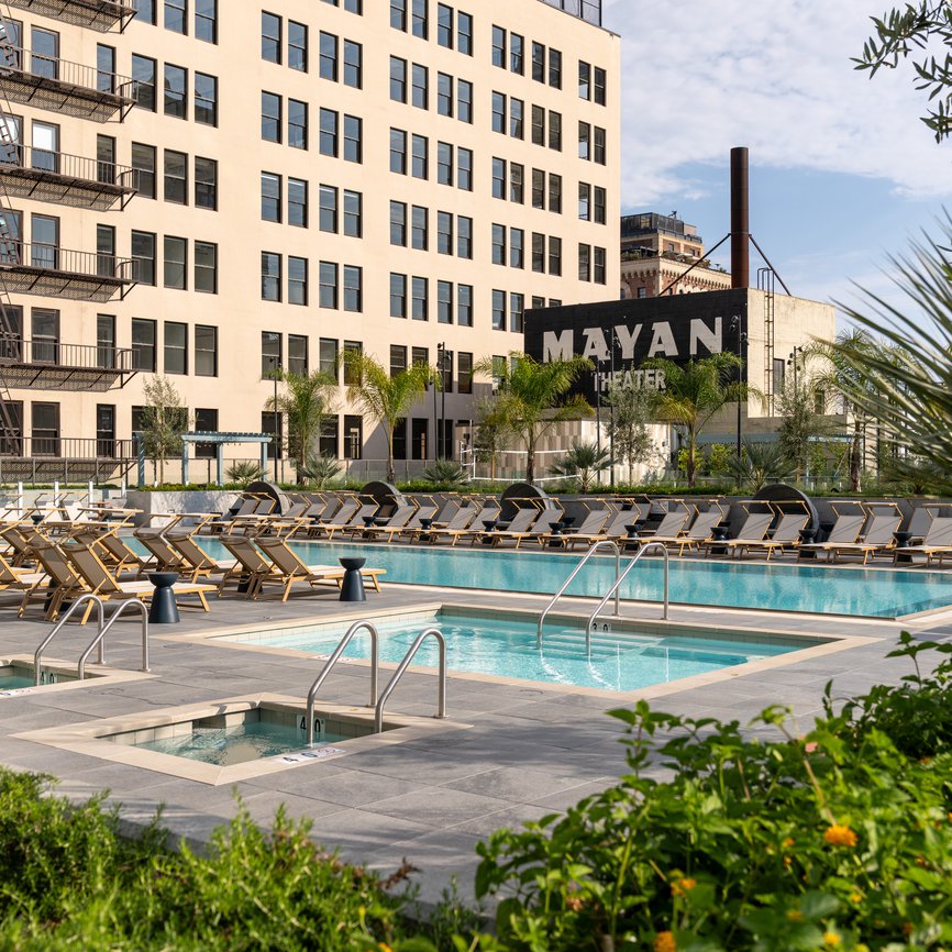 A modern outdoor pool area with lounge chairs and a hot tub, set in front of a beige multi-story building. In the background, a sign for the Mayan Theater is visible among palm trees and greenery.