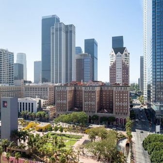 A view of downtown Los Angeles featuring tall modern skyscrapers, a historic brick building, and a green urban park with palm trees and walking paths in the foreground on a sunny day.