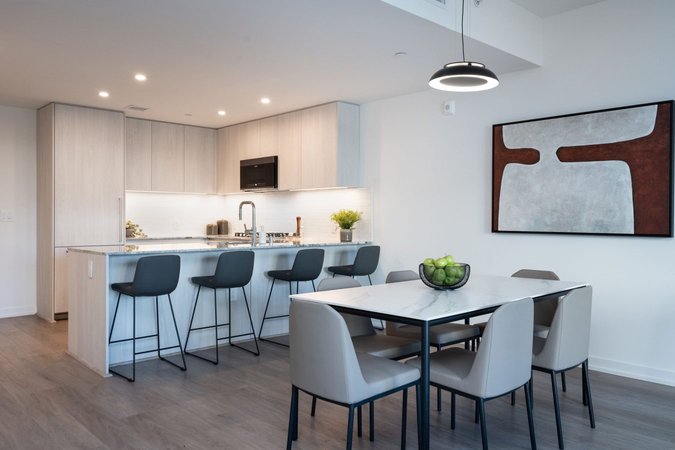 Modern kitchen and dining area with light wood cabinets, a large island with four gray bar stools, a dining table with six beige chairs, a bowl of green apples, and abstract wall art. Room is bright and neatly organized.