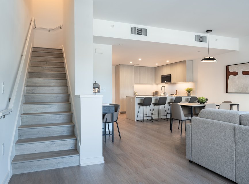 Modern open-concept apartment with light wood floors, a staircase on the left, a kitchen with light wood cabinets and a breakfast bar with stools, dining area, and a gray sofa in the foreground.
