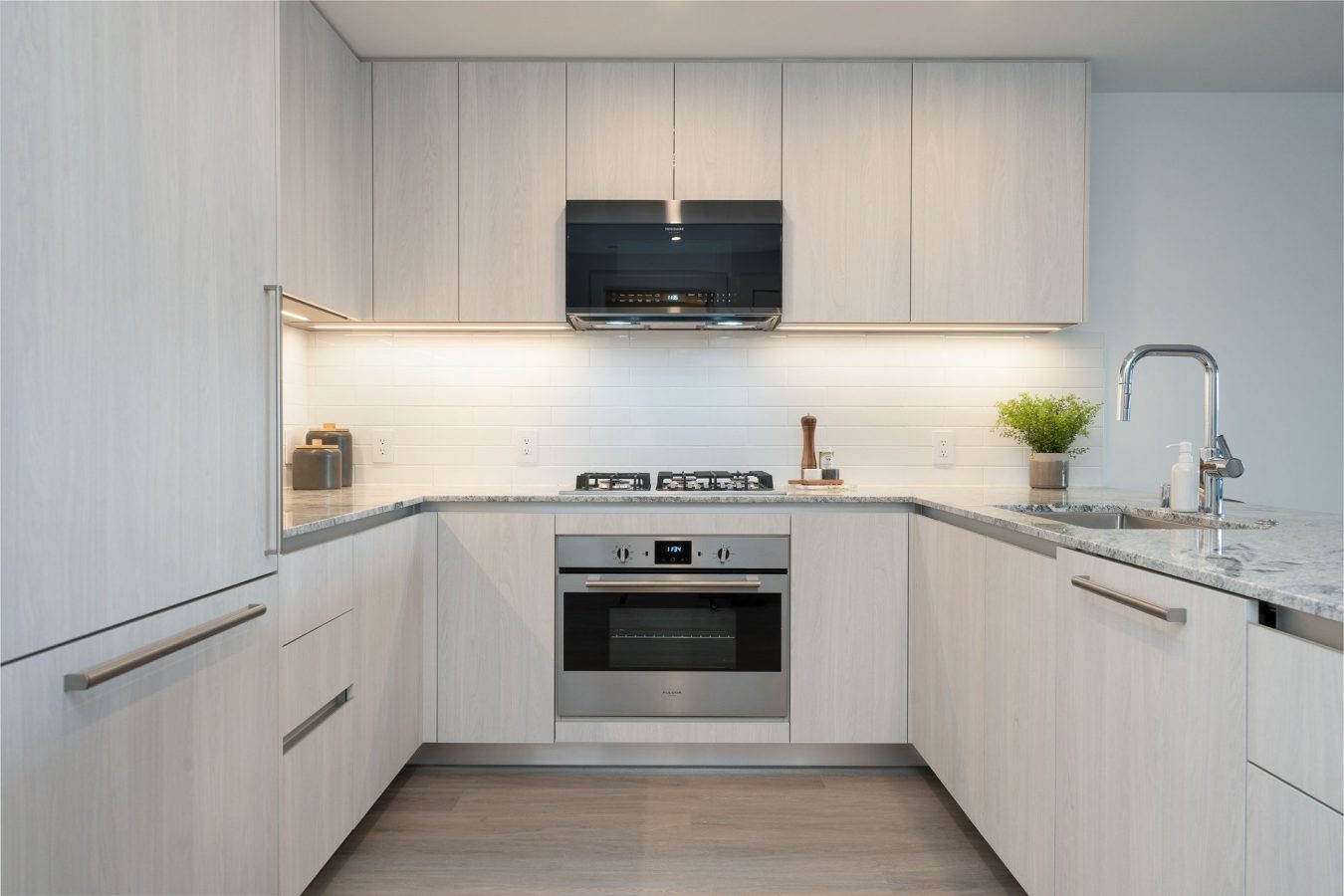 Modern kitchen with light wood cabinets, marble countertops, a built-in oven, gas stovetop, stainless steel microwave, sink with a faucet, and potted plant on the counter. The backsplash is white tile.