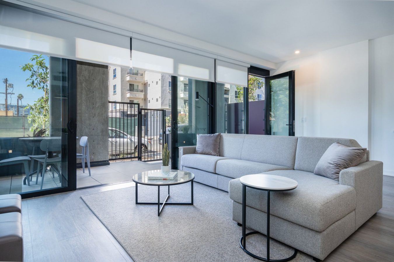Modern living room with a large light gray sectional sofa, coffee tables, and floor-to-ceiling glass doors leading to a small patio with outdoor seating and city views. The space is bright and minimalist.