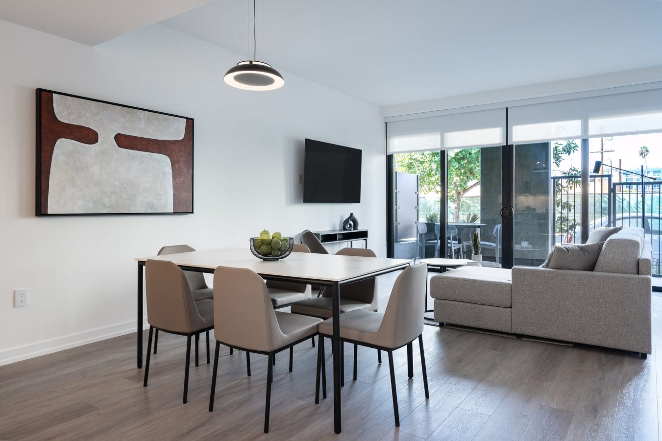 Modern dining and living area with a table, six beige chairs, wall art, a bowl of green apples, gray sectional sofa, mounted TV, and large windows with a view of a patio and outdoor greenery.