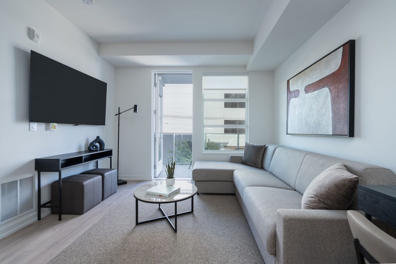 A modern living room with a large gray sectional sofa, abstract wall art, a round coffee table, a flat-screen TV mounted on the wall, and large windows letting in natural light.