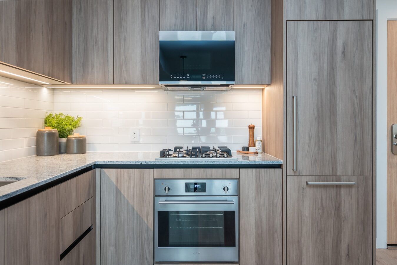 Modern kitchen with light wood cabinets, built-in stainless steel oven, gas cooktop, sleek range hood, white subway tile backsplash, and countertop with containers, a plant, and a pepper grinder.