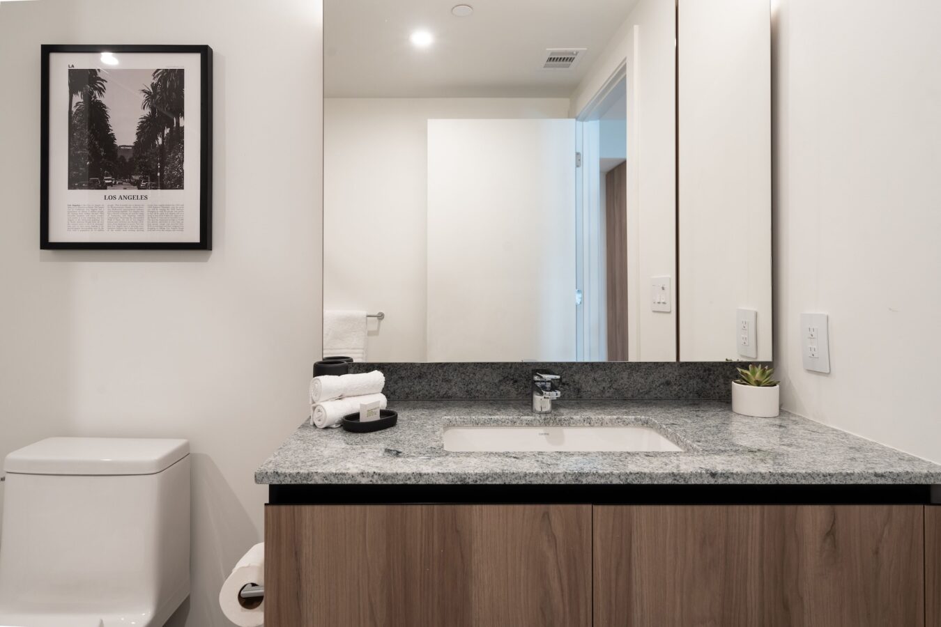 Modern bathroom with a gray countertop, wooden vanity, sink, small potted plant, rolled white towels, toilet, wall art of Los Angeles, and a large mirror reflecting the door and white walls.