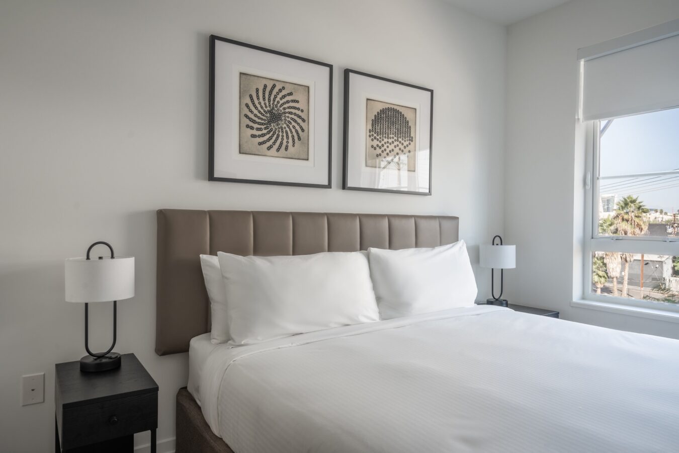 A modern bedroom with a neatly made bed, two white pillows, two bedside tables with lamps, and two abstract framed artworks above the bed. Sunlight comes in through a window with a partial city view.