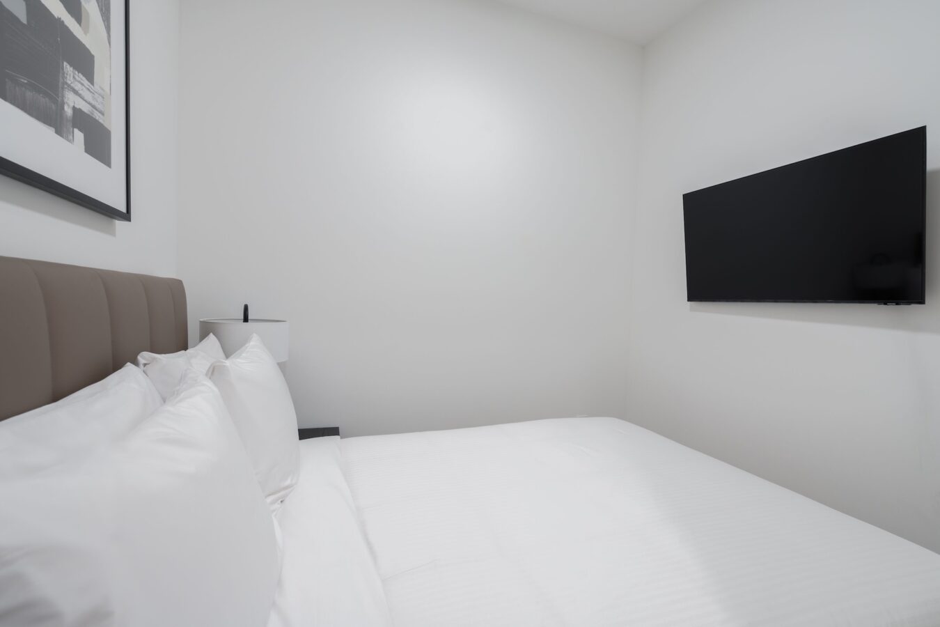 A minimalistic bedroom with a neatly made bed with white linens, a padded headboard, a wall-mounted TV, a white lamp, and a framed picture on the wall. The walls are painted white.