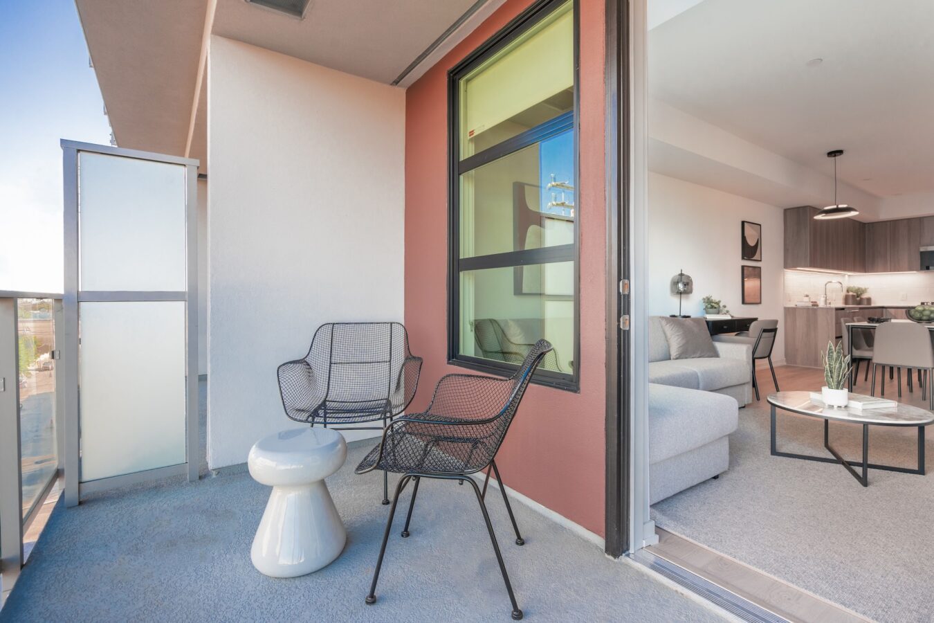 Modern apartment balcony with two black chairs and a small white table. The balcony overlooks a bright view and connects to a living room with a gray sofa, round coffee table, and open kitchen in the background.