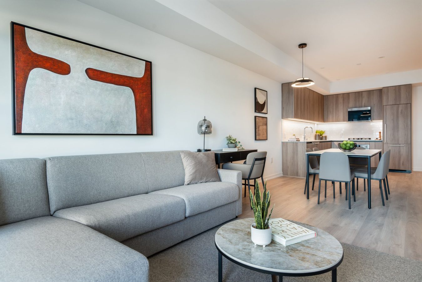Modern open-plan living space with a gray sofa, round coffee table with a plant and book, dining area, minimal decor, abstract art, and a kitchen with wood cabinets and pendant lighting in the background.