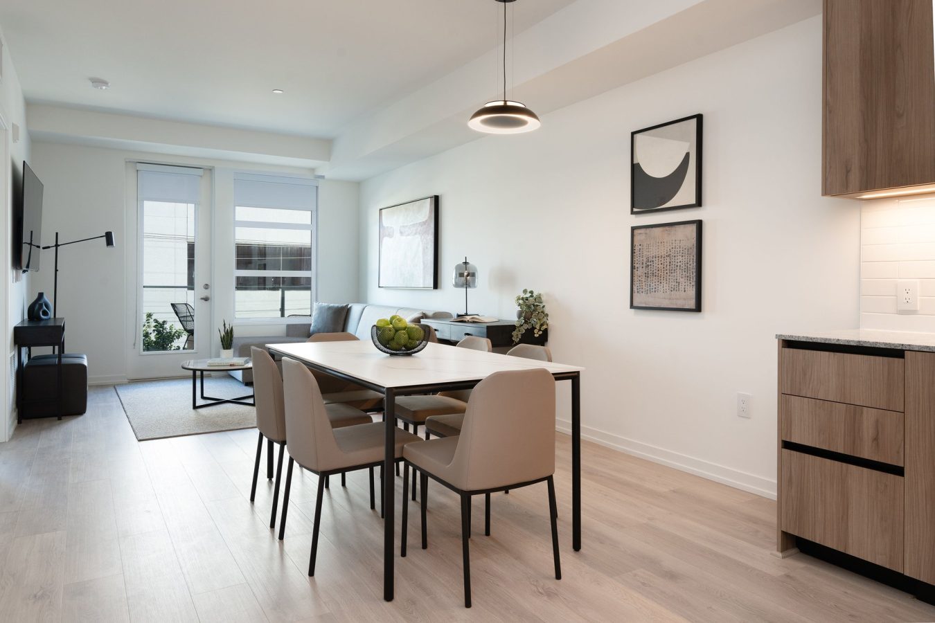 Modern, minimalist dining and living area with light wood floors, neutral-colored chairs around a table, wall art, and large windows letting in natural light. The kitchen is partially visible on the right.