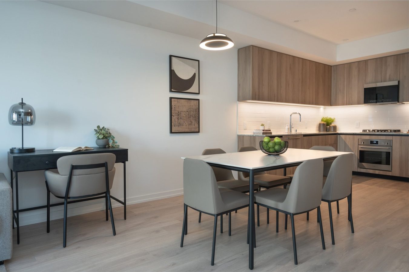 Modern kitchen and dining area with light wood floors, neutral chairs around a table with a bowl of green apples, a compact desk with a lamp and plant, and minimalist wall art, all lit by pendant lighting.