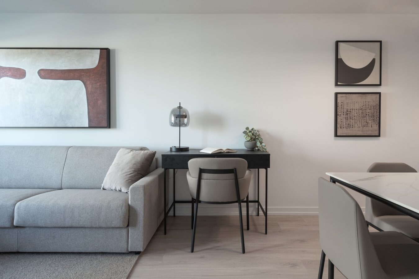 A modern living space with a light gray sofa, a black desk with a lamp and plant, neutral chairs, abstract wall art, and a minimal, clean aesthetic.
