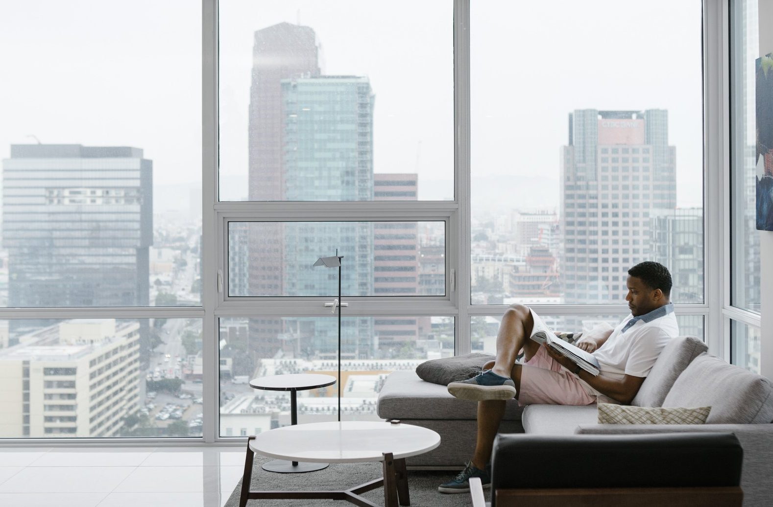 A man sits on a modern sofa reading a magazine in a bright, high-rise apartment with large windows, overlooking a cityscape of tall office buildings on a cloudy day.