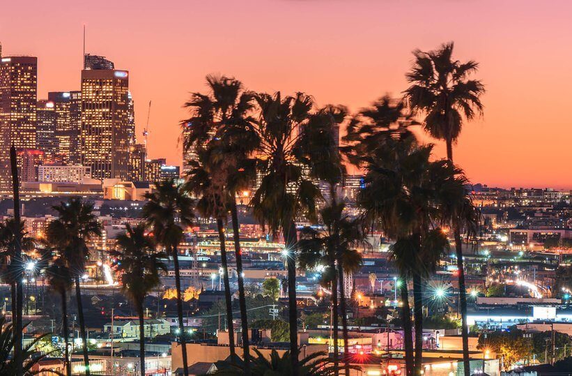 Cityscape at sunset with tall palm trees in the foreground, glowing city lights, and skyscrapers in the background under a pink-orange sky.