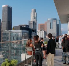 People gather and converse on a high-rise rooftop terrace overlooking a city skyline with tall modern buildings under a clear blue sky. Some guests lean on the glass railing, while others stand and chat in small groups.