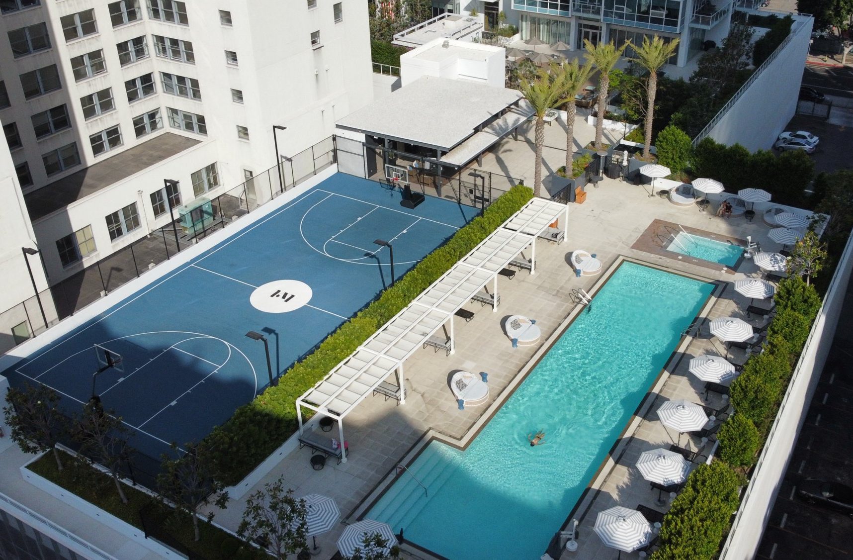 A rooftop view of a modern apartment complex features a blue basketball court, a rectangular swimming pool with a person swimming, lounge chairs, umbrellas, palm trees, and shaded seating areas.