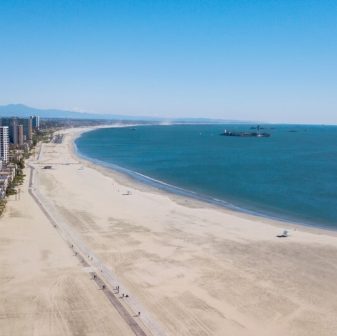 A wide, sandy beach stretches along the coastline under a clear blue sky, with calm ocean waves and a few distant ships. Tall buildings line one side of the beach, and a paved path runs parallel to the shore.
