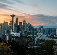 Seattle skyline at sunset with the Space Needle in the foreground, downtown skyscrapers, Mount Rainier in the background, and a mix of trees and buildings in the foreground.