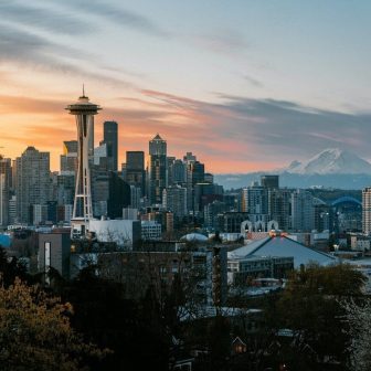 Seattle skyline at sunset with the Space Needle in the foreground, downtown skyscrapers, Mount Rainier in the background, and a mix of trees and buildings in the foreground.