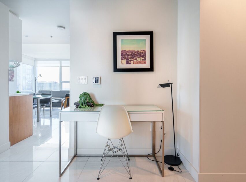 A modern, minimal home office with a white desk, white chair, desk lamp, a small plant, and a framed photo on the wall; bright natural light fills the space from large windows in the background.