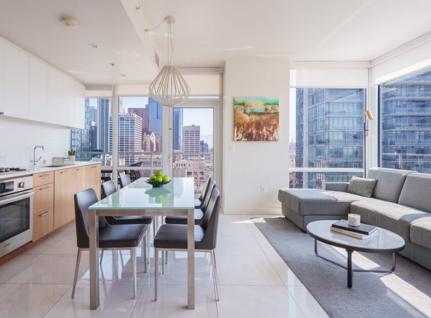 Modern apartment with large windows, city skyline views, a kitchen with light wood cabinets, a dining table with four chairs, a gray sectional sofa, and a colorful wall painting. Bright and airy atmosphere.