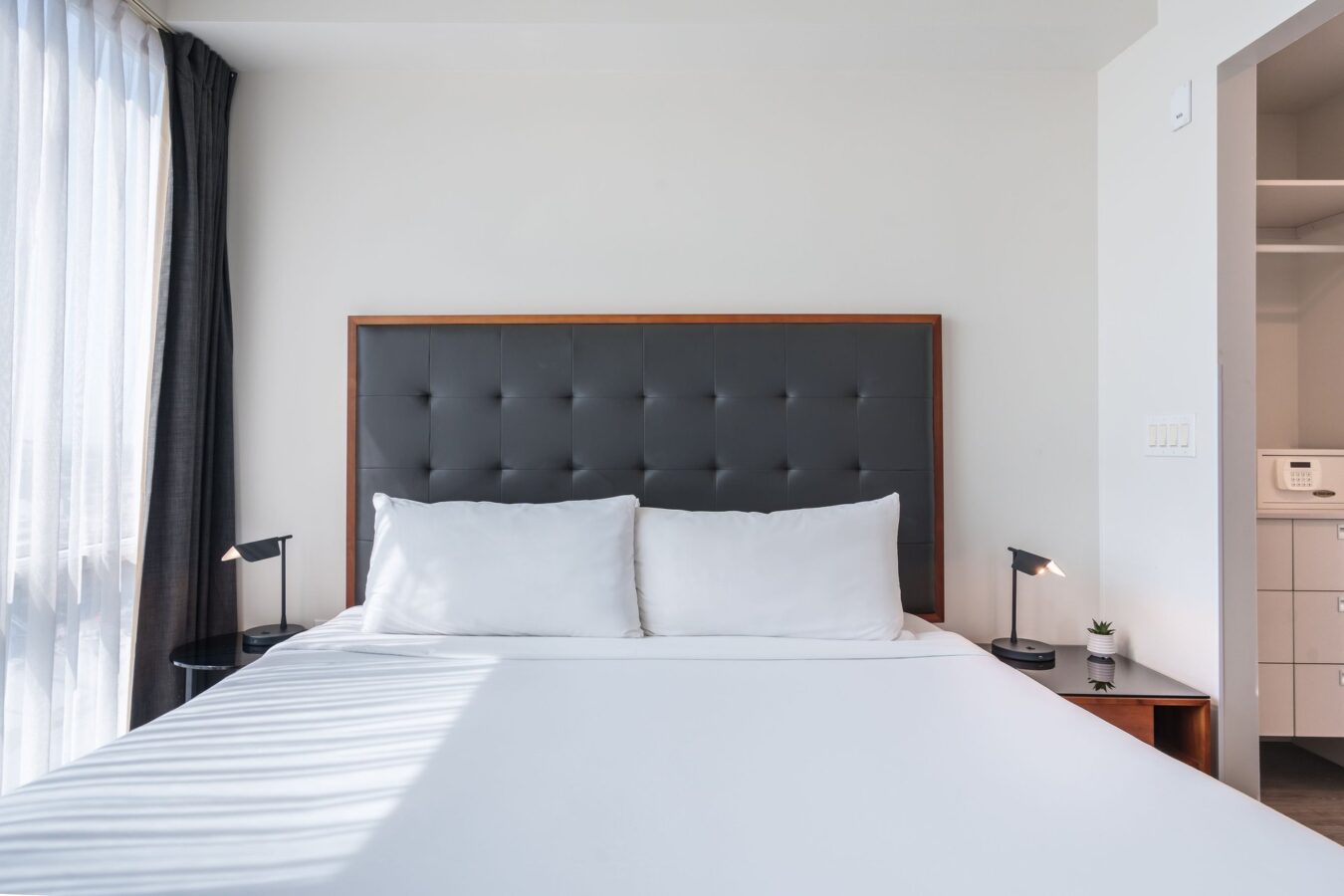 A modern, minimalistic bedroom with a neatly made bed, white bedding, a dark tufted headboard, two side tables with small lamps, and sunlight streaming through sheer curtains.