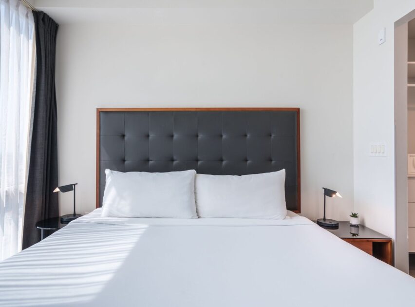A modern, minimalistic bedroom features a neatly made bed with white linens, a dark tufted headboard, two pillows, black bedside tables, matching lamps, and a small plant, with sunlight streaming in through a window.
