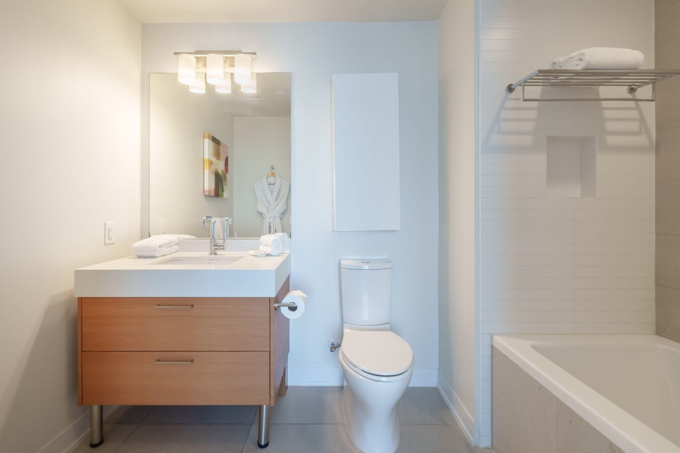 Modern bathroom with a wooden vanity, sink, mirror, and mounted light. A toilet is next to the vanity, and a bathtub with a towel rack is on the right. A bathrobe hangs on the wall in the background.