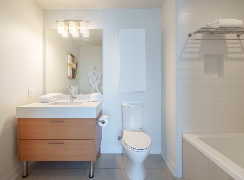 Modern bathroom with a wooden vanity, large mirror, toilet, bathtub, white towels, and a robe hanging on the wall. The space is bright, clean, and minimalistic with neutral tones.
