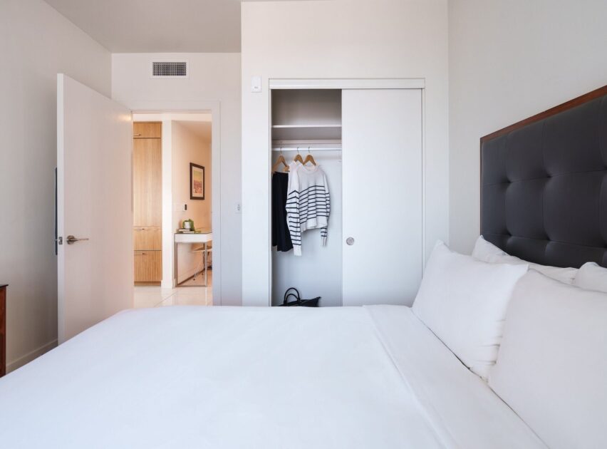 Minimalist bedroom with a neatly made bed, white linens, a closet with sliding doors, and a few clothes hanging inside. The door to the room is open, revealing a well-lit hallway and another room.