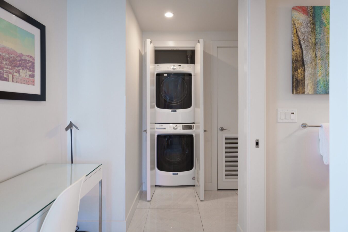 A compact laundry area with a stacked washer and dryer behind white double doors, located in a bright, modern room with a desk, chair, artwork, and towel rack visible.