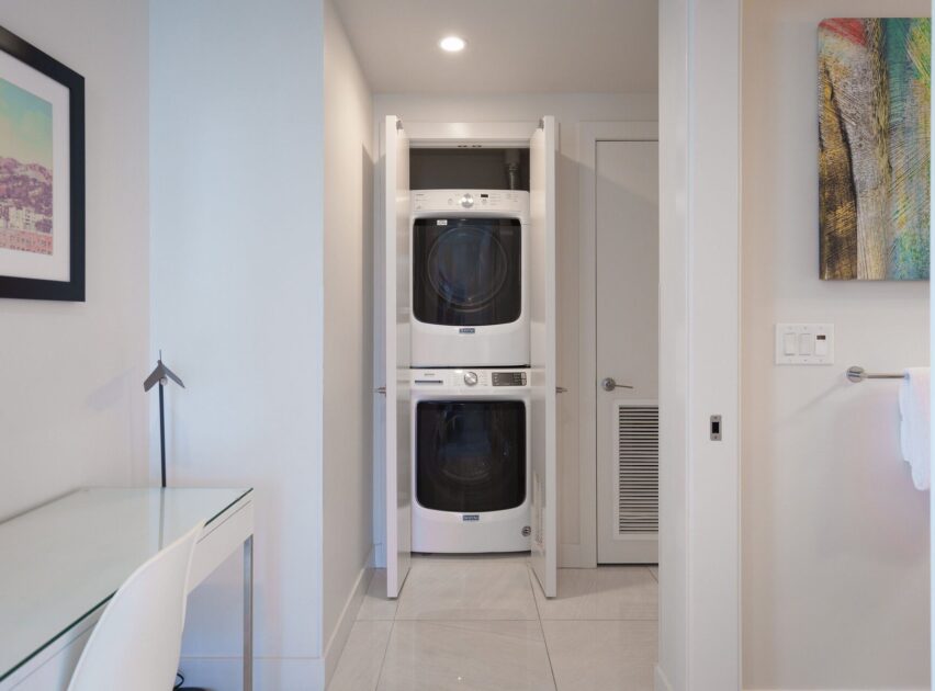 A compact laundry area with stacked washer and dryer behind open double doors in a modern, white room. Nearby are a desk with a white chair, framed art, and a towel hanging on the wall.